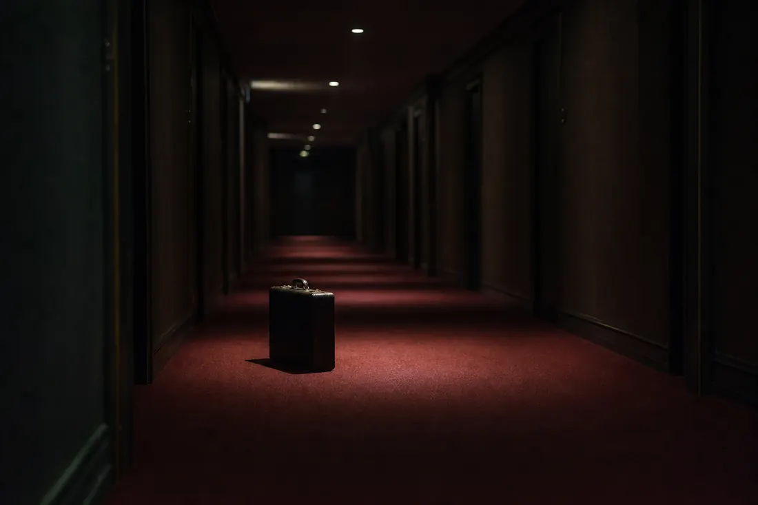 A dark hotel corridor with red carpet and one briefcase standing under a cone of light